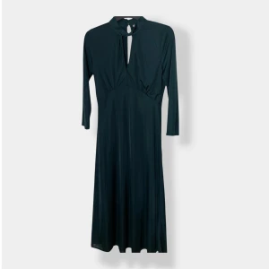 Mörkgrön långklänning med knyt - Snygg mörkgrön långklänning i glansigt material med trekvartsärm, hög hals och knyt fram. Klänningen har en cool slitsdetalj både fram och bak samt knappar i nacken. Perfekt för dig som gillar stilrena och unika plagg.