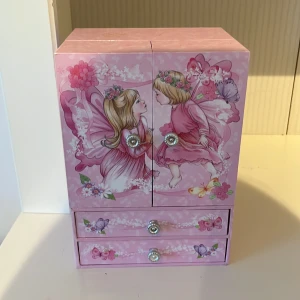 Rosa smyckeskrin med fe-motiv - Supergulligt rosa smyckeskrin med feer, blommor och fjärilar på utsidan. Skrinet har två dörrar, två lådor och en snurrande ballerina inuti. Perfekt för att förvara ringar, halsband och armband. Dekorativa knoppar i silverfärg och insida med blommigt mönster. Gör fin musik.