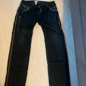 Mörkblå jeans från Diesel, stl 32 - Säljer ett par mörkblå jeans från Diesel i 100% bomull. Jeansen har snygga slitna detaljer vid fickorna, kontrastsömmar och raka ben. Klassisk femficksmodell med coola bakfickor och dragkedja i gylfen. Perfekta för dig som gillar en avslappnad stil.