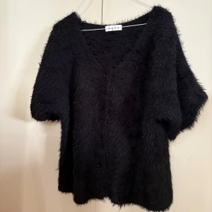 Svart fluffig kofta från Wera - Svart, kortärmad kofta från Wera med fluffig och mjuk känsla. Koftan har v-ringning, knappar framtill och en oversized passform. Perfekt för dig som gillar en chill och trendig look med lite extra textur.