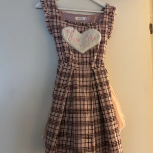 Rutig rosa klänning från Bobon21! - Supersöt kort rutig klänning i rosa och svart från Bobon21, Jnorri är ett märke som sålts hos Bobon21 som är en känd klädbutik i Japan!! Klänningen har volangdetaljer vid axlarna, ett fluffigt hjärta med broderad text på bröstet och knytband i midjan. Perfekt för dig som gillar gullig och unik stil! Det står storlek M men den är mer som en S, jag är 166cm och den är aningen kort på mig men den är perfekt att styla med en söt blus och ett par bloomers!!<3