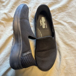 Svarta Skechers Slip-ins Arch Fit - Svarta Skechers Slip-ins med Arch Fit-sula och mesh-ovandel. Skorna är lätta att ta på tack vare slip-in-designen och har en tjock, bekväm sula. Perfekta för dig som vill ha maximal komfort och stöd i vardagen.