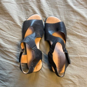 Svarta skinnsandaler från Remedios miralles - Svarta sandaler i skinn med coola utskurna detaljer och öppen tå. Sitter bekvämt med kardborreband runt vristen och har en platt sula med liten kilklack. Perfekta för varma dagar när du vill ha både stil och komfort.