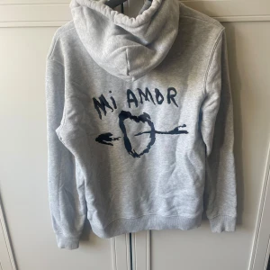 Grå hoodie med Mi Amor-tryck - Snygg grå hoodie med svart tryck på ryggen där det står 'Mi Amor', väldigt bra skick lite skrynklig på bilden därav den bara vart i garderoben men kan lätt fixas💕storlek S