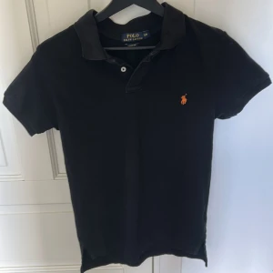 Svart pikétröja från Polo Ralph Lauren - Klassisk svart pikétröja från Polo Ralph Lauren med korta ärmar och orange broderad logga på bröstet. Tröjan har krage och två knappar vid halsen, skriv om funderingar.