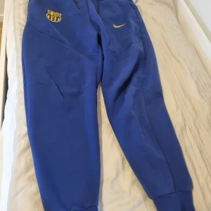 Blåa Nike FC Barcelona mjukisbyxor - Snygga blå mjukisbyxor från Nike med FC Barcelona-logga och gul swoosh på framsidan. Byxorna har dragsko i midjan och muddar vid bensluten. Perfekta för chill eller fotbollsträning. Mjuk och skön bomullsblandning.