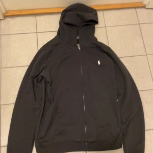 Svart hoodie från Polo Ralph Lauren - Svart hoodie från Polo Ralph Lauren i custom fit med dragkedja framtill och klassisk vit logga på bröstet. Tröjan har huva, långa ärmar och två fickor framtill. Perfekt för en chill och stilren look.