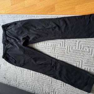 Svarta mjukisbyxor från Hugo Boss - Svarta mjukisbyxor från Hugo Boss med diskret logga på vänster ben. Byxorna har dragsko i midjan och är tillverkade i mjukt bomullsmaterial. Perfekta för chill dagar eller när du vill ha en clean och enkel look.