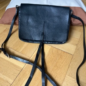 Svart axelväska i skinn från Wera - Stilren svart axelväska i äkta skinn från Wera. Väskan har långa remmar, lock med dekorativa band framtill och ett stort huvudfack med innerficka som stängs med dragkedja. Perfekt för dig som gillar minimalistisk design och snygga detaljer😍