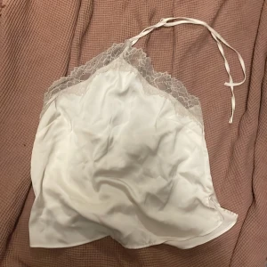 Vit satin tubtopp med spetsdetaljer - Snygg vit tubtopp i satin med spetskant upptill och tunna band som knyts i nacken. Toppen har en mjuk glansig yta och spetsen ger en romantisk vibe. Perfekt för dig som gillar en feminin och trendig look.