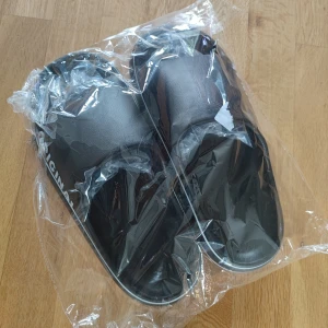 Svarta slides med vit text ORIGINAL - Svarta sandaler med bred rem och vit texten 'ORIGINAL' på sidan. Snygg och enkel design med vit rand längs sulan. Tillverkade i syntetmaterial och har öppen tå för en chill vibe. Perfekta för sommaren eller att glida runt hemma.