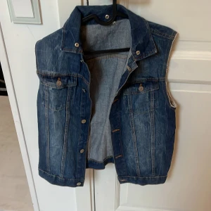 Klassisk blå jeansväst - Säljer en klassisk blå jeansväst med krage och knappar framtill. Västen har två bröstfickor med lock och knapp, samt snygga söm-detaljer. Perfekt att slänga över en hoodie eller t-shirt för en cool look.