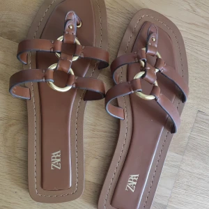 Bruna sandaler med gulddetaljer från Zara - Snygga bruna sandaler från Zara med platt sula och fyrkantig tå. Remmar i brunt skinnliknande material och coola guldfärgade ringdetaljer framtill. Perfekta för varma dagar och ger en trendig touch till din outfit.