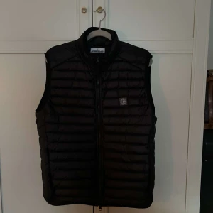 Svart dunväst från Stone Island - Säljer en svart dunväst från Stone Island med horisontella quiltade kanaler och dragkedja framtill. Västen har hög krage och en liten logotyp på bröstet. Tillverkad i återvunnen nylon och dunfylld för extra värme. Perfekt att slänga över en hoodie.