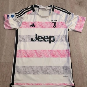 Juventus bortatröja 2023/2024 - Juventus officiella borta tröja från säsongen 2023/2024 med Jeep som sponsor. Tröjan är vit med svarta och rosa ränder, svarta detaljer och Serie A-märke på ärmen. Klassisk Adidas-logga och Juventus klubbmärke med tre stjärnor på bröstet.
