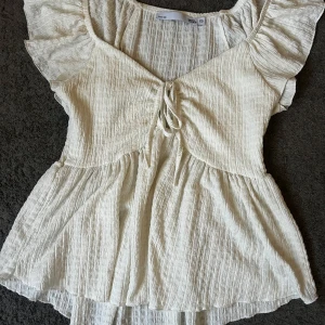 Krämvit blus med volangärm från lager 157 - Supersöt krämvit blus från lager 157 i storlek XS. Blusen har volangärmar, v-ringning och knytning framtill. Tillverkad i ett lätt, strukturerat bomullstyg som ger en luftig känsla. Perfekt för varma sommardagar. Använd ett fåtal gånger, säljer då den är för stor för mig💞