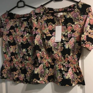 2 st Blommig topp från Adele & Co strl 122 - Supersöt blommig topp från Adele & Co i 100% bomull. Svart botten med färgglada blommor i lila, rosa, gul och grön. Kortärmad modell med smock i midjan och volangkant nedtill.  149kr styck är Ordinariepris.. Obs storlek 122