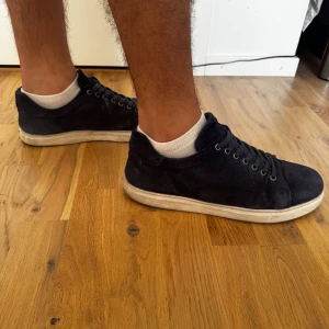 SANDÖ Sneakers - Snygga mörkblå sneakers i mocka med klassisk rund tå och platt vit sula. Skorna har svarta skosnören och metallöljetter för en stilren look. Perfekta för dig som gillar en clean och enkel stil med lite extra känsla tack vare mockamaterialet.