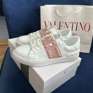 Rosa Valentino skor - Snygga Valentino Garavani Rockstud sneakers i vitt skinn med ljusrosa band och ikoniska nitar. Klassisk rund tå, platt sula och vita snören. Perfekt för dig som vill sticka ut med exklusiva detaljer och modern design.