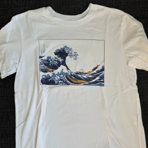Vit t-shirt med vågtryck från Even&Odd - Vit t-shirt från Even&Odd med ikoniskt tryck av Hokusais stora våg i blått och gult på bröstet. Klassisk rund hals och korta ärmar. Tillverkad i mjuk bomull, perfekt för en chill och stilren look.