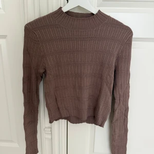 Brun ribbad stickad tröja - Säljer en brun ribbad stickad tröja med croppad passform och hög hals. Tröjan har långa ärmar och ett snyggt mönster med breda ribbor som ger en modern look. Perfekt att styla med jeans eller kjol för en chill vibe.