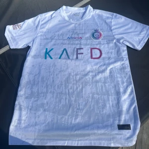 Al Nassr Ronaldo 7 vit fotbollströja - Snygg vit Al Nassr fotbollströja med Ronaldo 7 på ryggen. Tröjan är i storlek M och har används fåtal gånger.