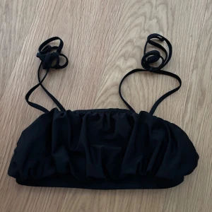 Svart bikiniöverdel med snörning M - Svart bikiniöverdel i bandeau-modell med rynkade detaljer och tunna axelband som knyts i nacken. Snygg och enkel design som passar perfekt till sommaren och strandhäng. Tillverkad i stretchigt syntetmaterial för skön passform. ser ny ut!