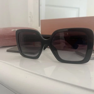 Svarta solglasögon från Miu Miu - Snygga svarta solglasögon från Miu Miu med fyrkantig oversized båge och mörka glas. Sidorna har guldig Miu Miu-logga som ger en lyxig vibe. Kommer med originalfodral och putsduk. Perfekta för dig som vill sticka ut med en statement-accessoar.