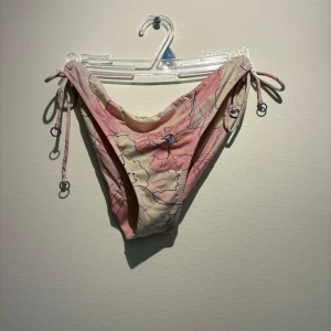 Blommig bikini från Change XS - Supersöt bikini från Change med blommigt mönster i ljusrosa och beige toner. Modellen har knyt i sidorna med små ringdetaljer och en feminin, lite högre skärning. Perfekt för strandhäng och poolparty!