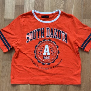 Orange cropped T shirt South Dakota från Sin say  - Ny stilig T shirt med print South Dakota National Ecological Academy 1891 i mitten. Med design på armar också. Cropped och wide t shirt som ger en modern och avslappnad look. Köptes från Sin Say. 
