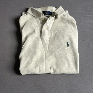 Ljusgrå pikétröja från Ralph Lauren - Snygg ljusgrå pikétröja från Ralph Lauren med klassisk krage och två knappar framtill. Ikoniska gröna polospelaren broderad på bröstet. Tröjan är i bomull och har korta ärmar, perfekt för en clean och stilren look.