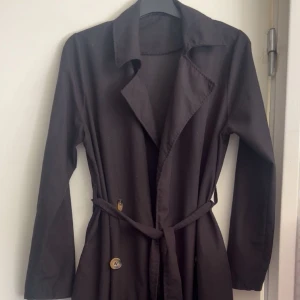 Svart trenchcoat med bälte - Snygg svart trenchcoat med klassisk krage, knappar framtill och knytskärp i midjan. Lång modell med raka ärmar och stilren look, perfekt för dig som gillar minimalistisk stil. Jackan har en tidlös design som funkar till många outfits.