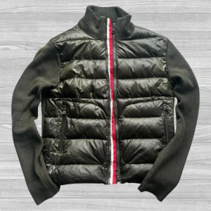 Moncler Cardigan - Snygg olivgrön cardigan från Moncler i storlek M. Jackan har stickade ärmar och krage, quiltad front i glansigt material och dragkedja med röd och vit detalj. Perfekt för dig som vill ha en stilren och sportig look.