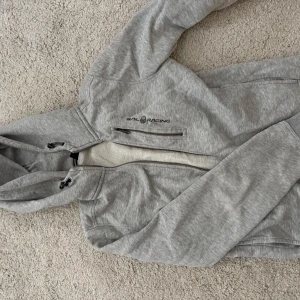 Grå hoodiejacka från Sail Racing - Mysig grå hoodiejacka från Sail Racing med dragkedja och huva. Jackan har mjukt fleecefoder på insidan, praktiska fickor med dragkedja och justerbar dragsko i huvan. Perfekt för kyliga dagar när du vill vara både bekväm och snygg.