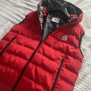 Röd dunväst från Moncler med huva - Snygg röd dunväst från Moncler med svart innerfoder och avtagbar huva. Västen har dragkedja framtill, två fickor med dragkedja och Moncler-logga på bröstet. Stora Moncler-tryck på huvkanten ger en extra cool touch. Har använd den ba tre gånger passar inte mig längre 