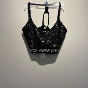 Nike Pro svart och guld linnetopp - Nike Pro linnetopp i svart med guldiga blommönster och små prickar. Snygg sportig design med bred resår med logga och cool rygg med banddetaljer. Perfekt för träning eller när du vill ha en statement-top.
