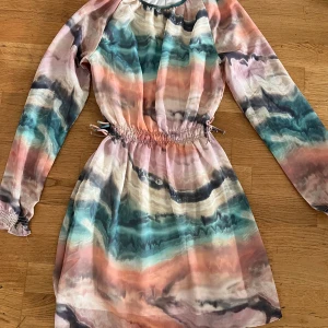 Färgglad kortklänning med långa ärmar - Superfin kortklänning med långa ärmar och elastisk midja. Klänningen har ett unikt mönster i pastelliga toner av rosa, blått, beige och svart. Materialet är tunt och flowy, perfekt för varmare dagar. Snygg och bekväm med coola färgskiftningar.