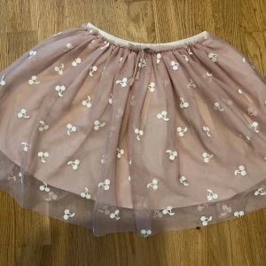 Rosa tyllkjol med körsbär från H&M - Supersöt rosa kortkjol från H&M i tyll med vita broderade körsbär och glittrig resår i midjan. Kjolen har ett tunt, luftigt lager och en mjuk underkjol. Perfekt för dig som vill ha en lekfull och gullig stil.