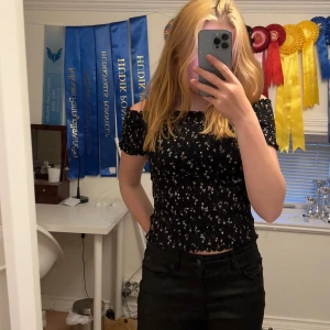 Svart smockad blommig offshoulder topp - Supersöt svart smockad topp med små blommor i vitt och rosa. Offshoulder-modell med korta ärmar och croppad passform. Perfekt för sommarens alla häng! Toppens material är stretchigt och sitter tajt men bekvämt.