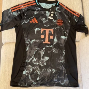 FC Bayern München Adidas fotbollströja - Snygg FC Bayern München fotbollströja från Adidas med svart bas och marmorerat mönster i ljusblått. Tröjan har korta ärmar, orange detaljer och klubbmärke på bröstet. Tillverkad i lätt och ventilerande material, perfekt för träning eller match.