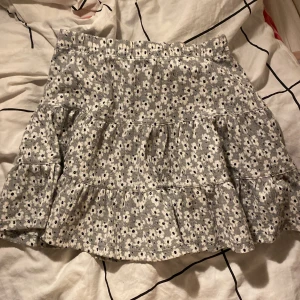 grön blommig volangkjol - Supersöt kort kjol i gönt med små vita blommor och mörka detaljer. Kjolen har volanger och en avslappnad passform, perfekt för en chill och trendig look. Materialet känns mjukt och lätt, vilket gör den skön att bära under varmare dagar.