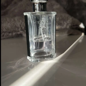 Prada L'Homme Eau de Toilette - Snygg parfymflaska från Prada med stilren design i genomskinligt glas och svarta detaljer på sidorna. Flaskans form är kantig och modern, med silverfärgat sprayhuvud. Perfekt för dig som gillar exklusiva dofter och vill ha något lyxigt på hyllan.