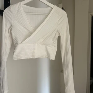 Vit omlottopp med ribbad struktur - Snygg croppad omlottopp i vitt med ribbad struktur och långa ärmar. Toppen har en v-ringad omlottdesign som ger en stilren och trendig look. 