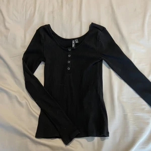 Svart ribbad långärmad topp Pieces - Svart långärmad topp från Pieces med ribbad struktur och knappdetaljer framtill. Snygg, figurnära passform och rundad halsringning. Perfekt att styla med jeans eller kjol för en clean look.