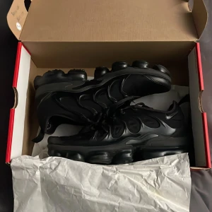 Svarta Nike Air Vapormax sneakers - Säljer ett par helsvarta Nike Air Vapormax sneakers med futuristisk design och synlig dämpning i sulan. Skorna har snörning, rund tå och är tillverkade i syntetmaterial med coola detaljer på ovandelen. Perfekta för dig som gillar streetstyle och vill ha något unikt på fötterna.