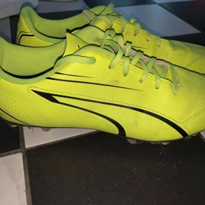 Neongula fotbollsskor från Puma - Snygga neongula fotbollsskor från Puma med svarta detaljer och klassisk snörning. Skorna har en smal passform och är tillverkade i syntetmaterial, perfekta för dig som vill sticka ut på planen.