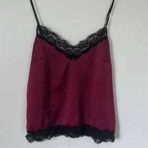 Vinröd linnetopp med spetsdetaljer - Snygg vinröd linnetopp med tunna axelband och svarta spetsdetaljer längs urringning och nederkant. Toppen har en v-ringad form och är perfekt för dig som gillar en chic och feminin stil.