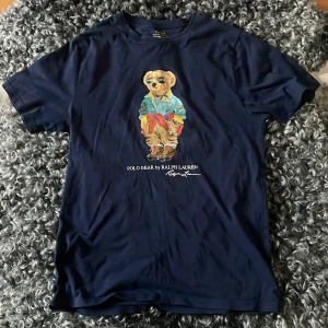 Mörkblå Polo Bear t-shirt Ralph Lauren - Snygg mörkblå t-shirt från Ralph Lauren med ikoniska Polo Bear-trycket på bröstet. Klassisk passform, korta ärmar och rund halsringning. Tillverkad i mjuk bomull som känns skön mot huden. (Storleken är 170 barnstorlek men den sitter som S i passform)