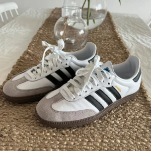 Adidas Samba OG vit/svart 39 1/3 - Adidas Samba OG sneakers i vitt skinn med grå mockadetaljer och svarta ränder. Klassisk gummisula och guldfärgad Samba-text på sidan. Snygg retrostil med snörning och rund tå, perfekt till streetwear eller casual outfits. Helt oanvända! Lappen tog jag av innan jag märkte att skorna inte passade mig. Säljer för att kunna köpa nya i min storlek. Köparen står för frakten. 