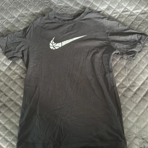 Svart Nike t-shirt med basketlogga - Svart t-shirt från Nike med klassisk rund hals och korta ärmar. På bröstet finns en stor vit Nike Swoosh-logga med basketbollmönster. Skön passform och tillverkad i mjuk bomull, perfekt för dig som gillar sportig stil.